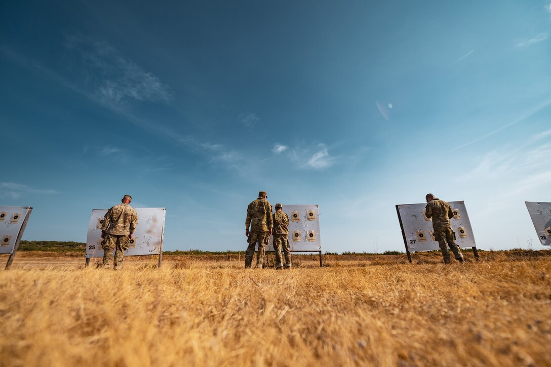 Soldiers check shooting targets in a field under a blue sky.