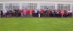A group of over 50 people in civilian cloths stand on green grass in front of a white wall with windows.