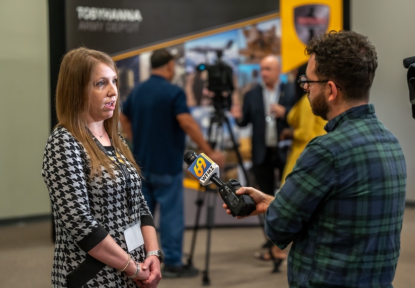 A person speaks to a news reporter. The reporter is holding a microphone. In the background, another person is being interviewed on camera.