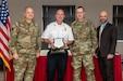 The image depicts four people posing for a photo during an awards ceremony.  Two are soldiers in military uniforms, one is wearing a suit, and the other is wearing black pants and a white shirt denoting his position as Chief of a Fire unit.  He is holding a plaque.