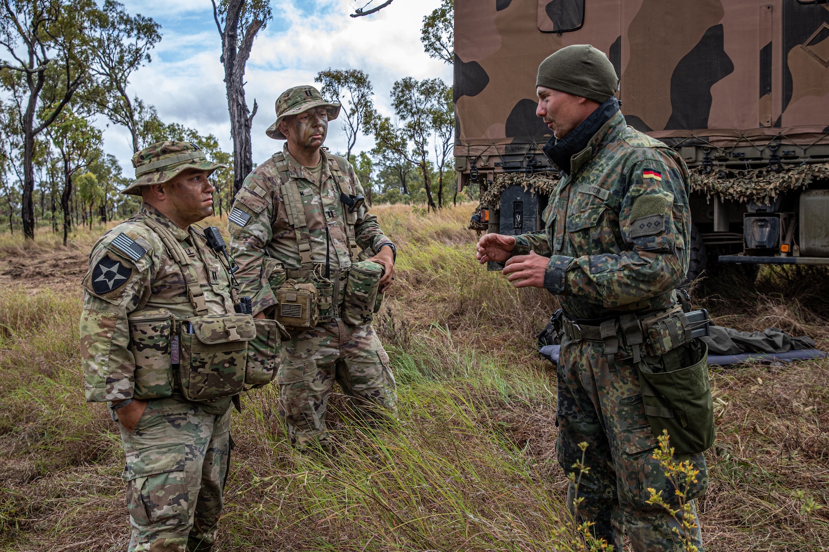 military men in a field, talking, with a vehicle behind them.