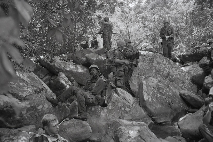 About a half-dozen men in uniform relax on rocks while holding rifles.