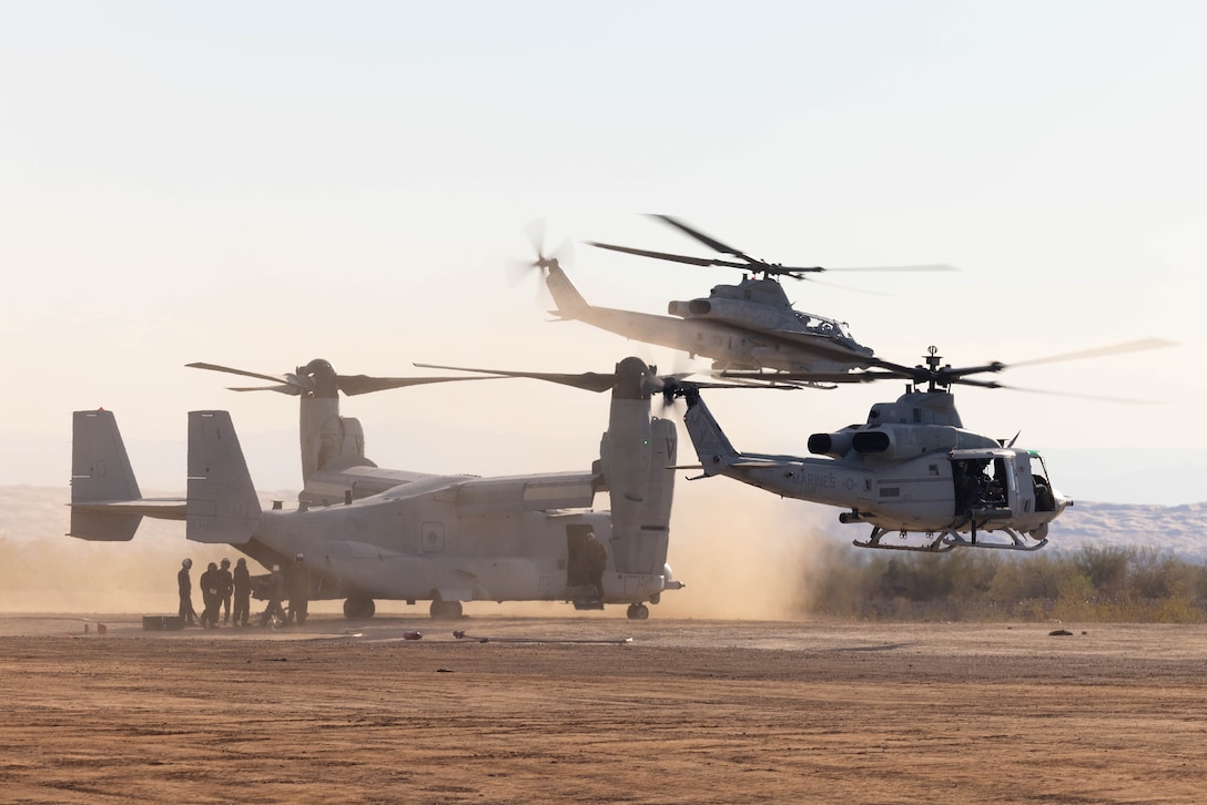 A military aircraft is parked on a desert area as two other aircraft hover nearby during daylight.