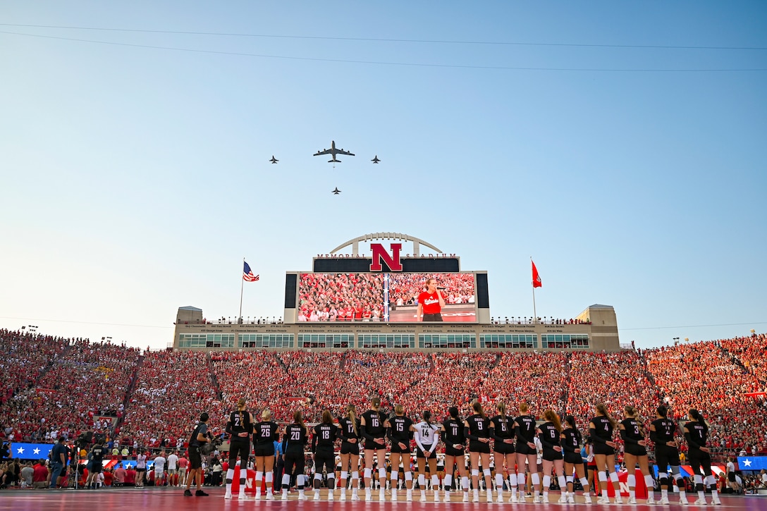 Volleyball players stand in formation facing a crowd standing in bleachers at a stadium as four aircraft fly above.