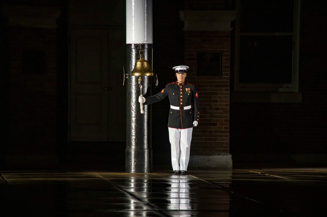 A Marine wearing a ceremonial uniform rings a bell while standing at attention in a dark area.