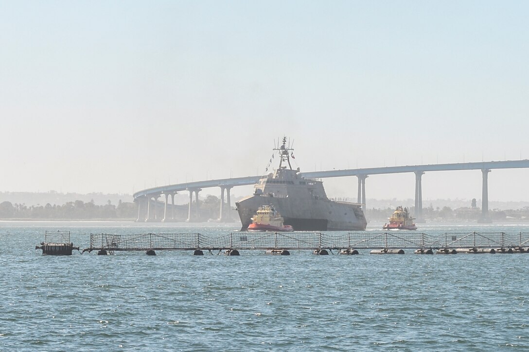 A ship transits a body of water next to two boats with a bridge in the background and netted fence in the foreground.