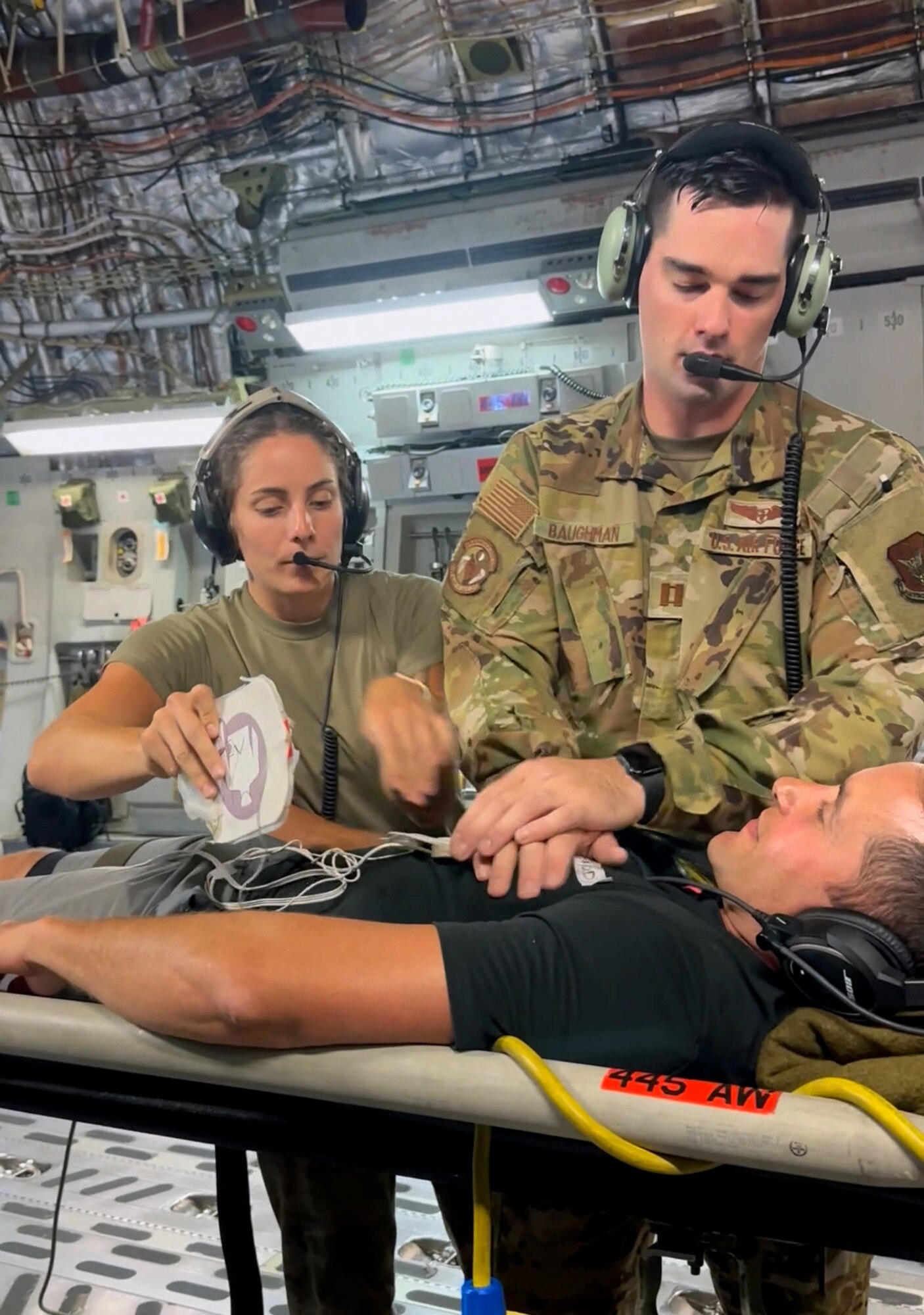 U.S. Air Force Capt. Kyle Baughman, right, 445th Aeromedical Evacuation Squadron flight nurse, provides CPR to mock patient Matt O’Hara, DynastyNerds podcast, as Senior Airman Esa Edwards, 445th AES flight nurse, prepares to roll O’Hara over to apply adhesive defibrillator pads per the training scenario, on board a 445th Airlift Wing C-17 Globemaster III aircraft, Aug. 23, 2023. O’Hara and fellow DynastyNerds podcasters visited with the wing to learn about the air evacuation mission and also participated as moulage patients to help the AES get hands-on training with live “patients.” (U.S. Air Force photo by 445th Airlift Wing Public Affairs)(Photo taken with phone)