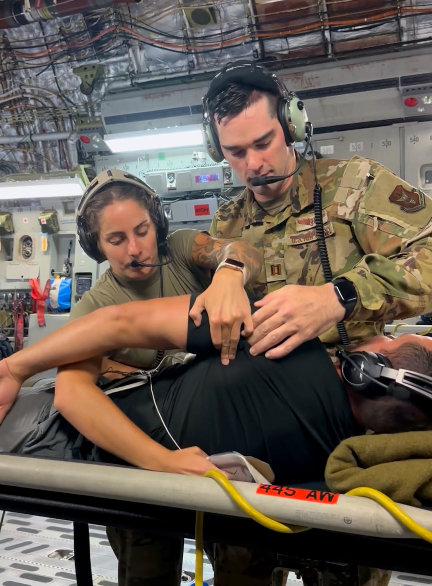 U.S. Air Force Capt. Kyle Baughman, right, 445th Aeromedical Evacuation Squadron flight nurse, and  Senior Airman Esa Edwards, 445th AES flight nurse, roll over mock patient Matt O’Hara, DynastyNerds podcast, to apply adhesive defibrillator pads per the training scenario, on board a 445th Airlift Wing C-17 Globemaster III aircraft, Aug. 23, 2023. O’Hara and fellow DynastyNerds podcasters visited with the wing to learn about the air evacuation mission and also participated as moulage patients to help the AES get hands-on training with live “patients.” (U.S. Air Force photo by 445th Airlift Wing Public Affairs)(Photo taken with phone)