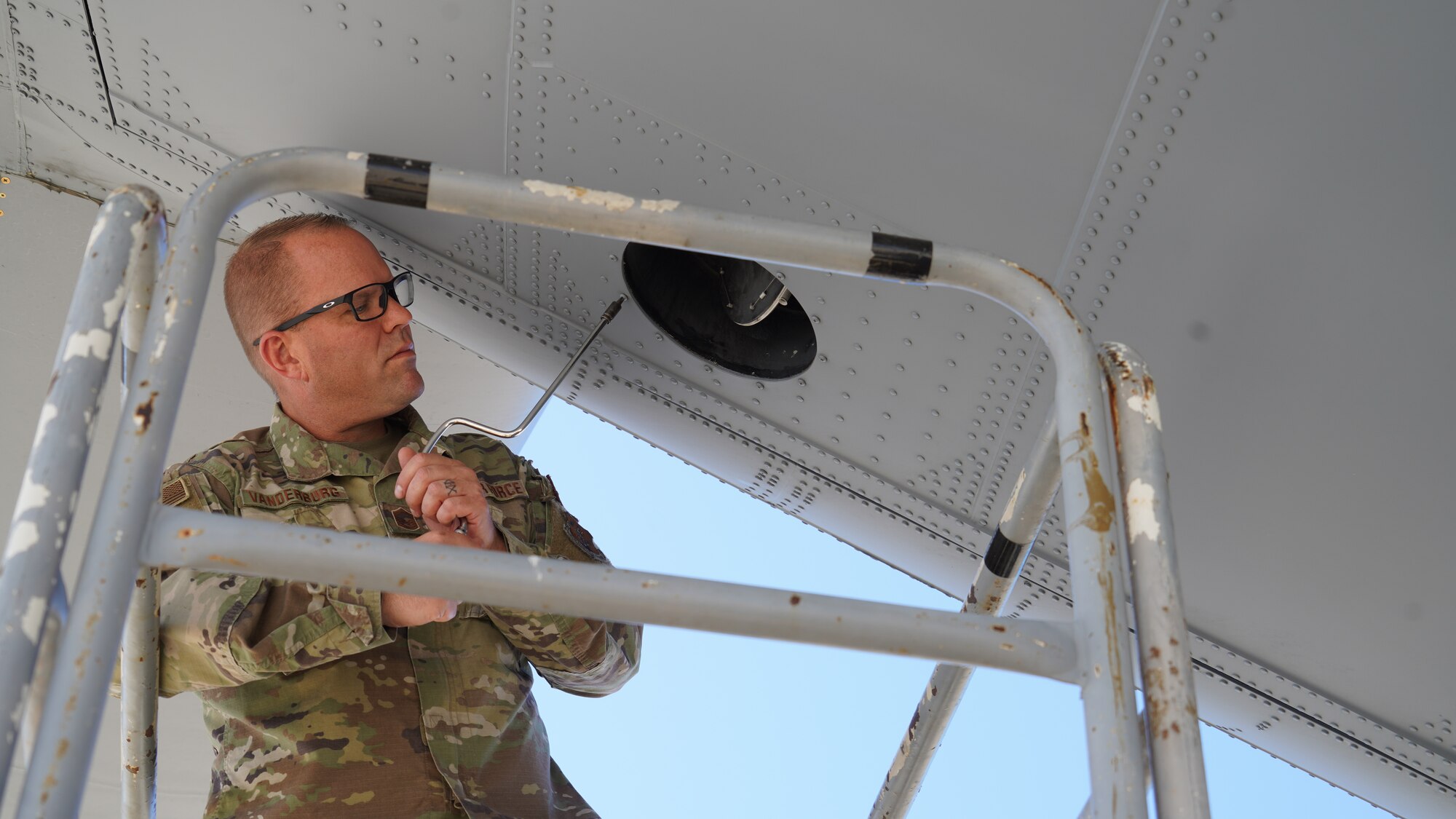 A photograph of a California Air National Guard Airman / Maintainer on top of an aircraft ladder using a specialized elongated tool to remove a bolt from behind and underneath the tail of a C-130J Super Hercules.