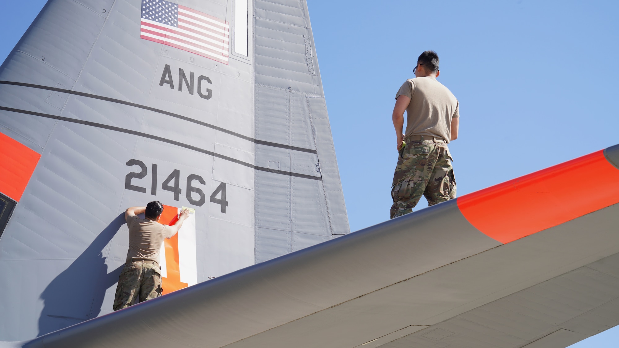 A photo of two California Air National Guard Airmen standing on the on top of a C-130J Super Hercules as they add a bright orange numbered decal to the flash near the back of the Aircraft.