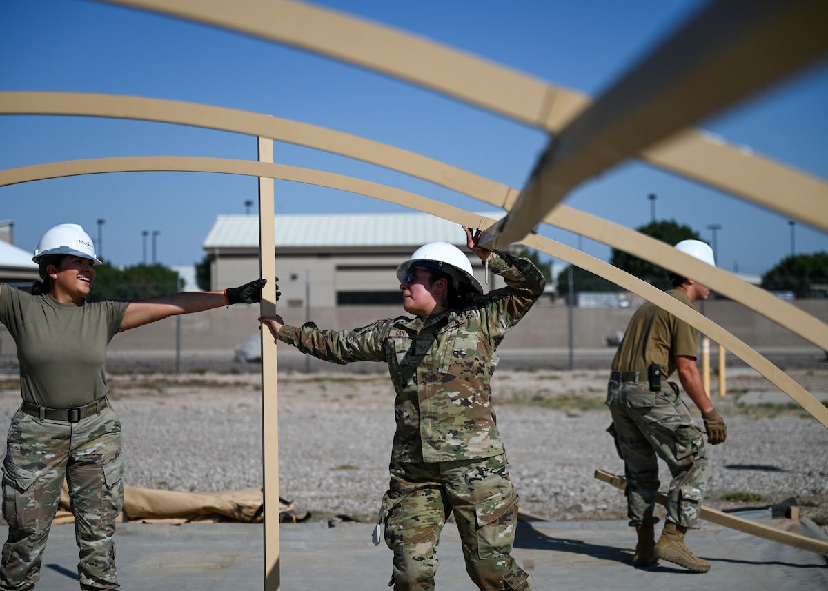 A photo of people in military uniforms assembling the metal structure of a large tent.