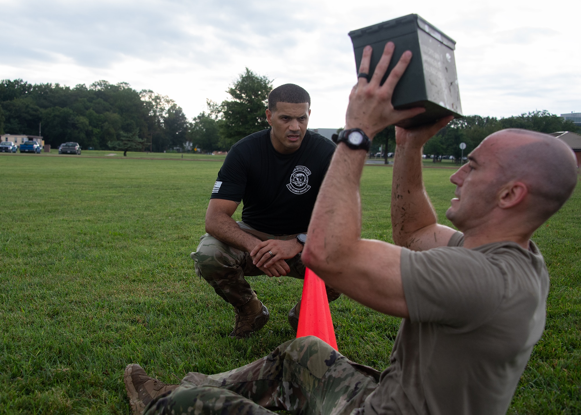 A man encourages another man while he lifts an ammo can over his head.
