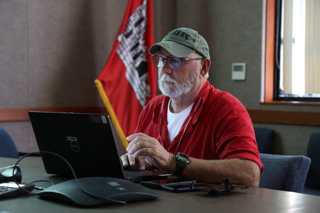 A man in a baseball cap and red polo shirt looks toward a laptop computer screen; the U.S. Army Corps of Engineers flag appears in the background.