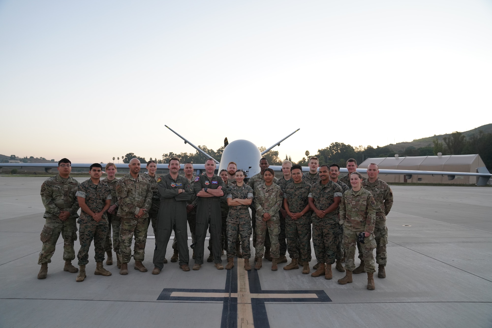 A group of people in Air Force and Marine Corps uniforms stand in front of an unmanned aircraft.