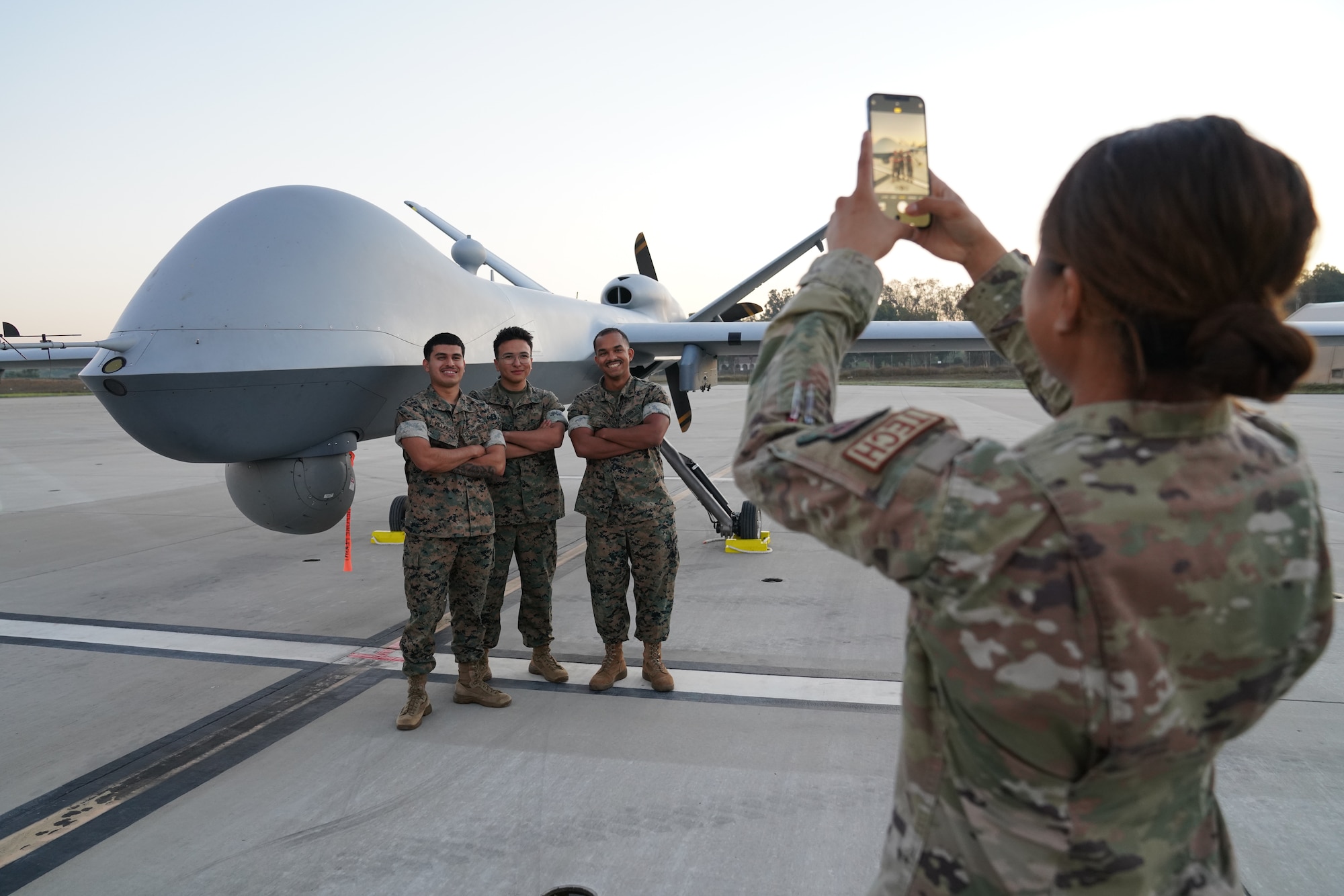 A woman in a military uniform takes a picture of three men in military uniforms in front of an unmanned aircraft.