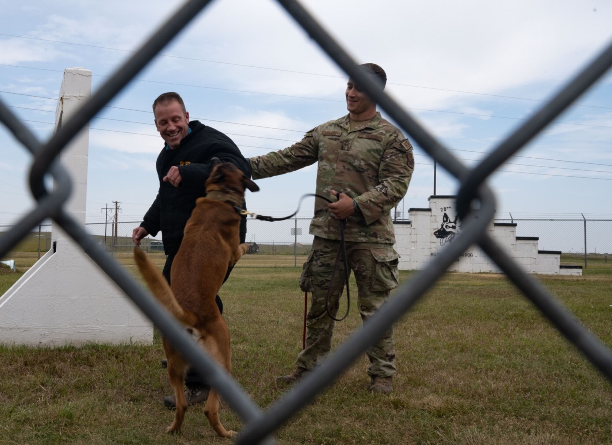 Aron-D405, 28th Security Forces Squadron Military Working Dog, (center) demonstrates the technique he would use to take down a bad guy on Mike Pogany, 28th Operations Support Squadron honorary commander, left, with his partner, Staff Sgt. John Cammarata, 28th SFS Military Working Dog kennel master, during a tour for Honorary Commanders at Ellsworth Air Force Base, South Dakota, Aug. 25, 2023.