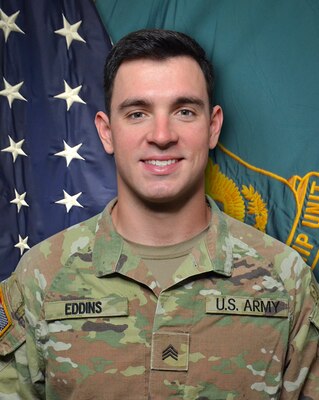 Man in U.S. Army uniform standing in front of two flags.