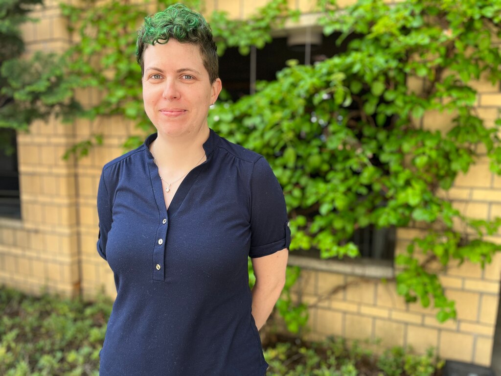 A woman in a polo-style shirt stands in front of a brick wall and a tree.