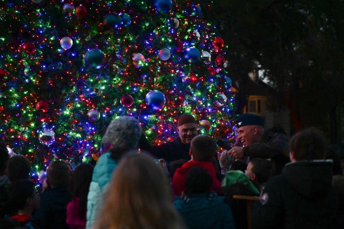 Many Children gather around a button that lights up a Christmas tree which fills up the background