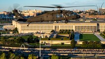 An Army helicopter is flying in front of the Pentagon, and there are thousands of people filling one of the roadways below. In the distance is the three silver spires of the Air Force Memorial.