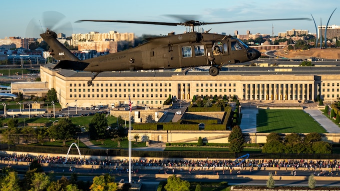 An Army helicopter is flying in front of the Pentagon, and there are thousands of people filling one of the roadways below. In the distance is the three silver spires of the Air Force Memorial.