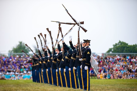 Army soldiers are standing in a row on a grass field. Some have tossed rifles into the air, and others are holding their rifles upside down in a contagion of movement.