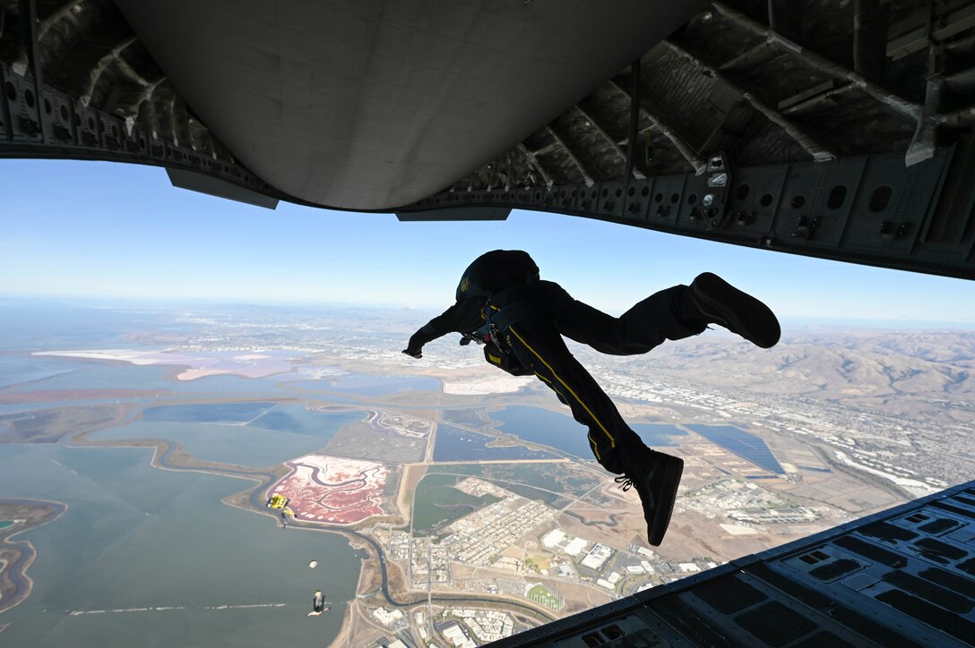A service member jumps from an aircraft during daylight. Water and land can be seen in the distance.