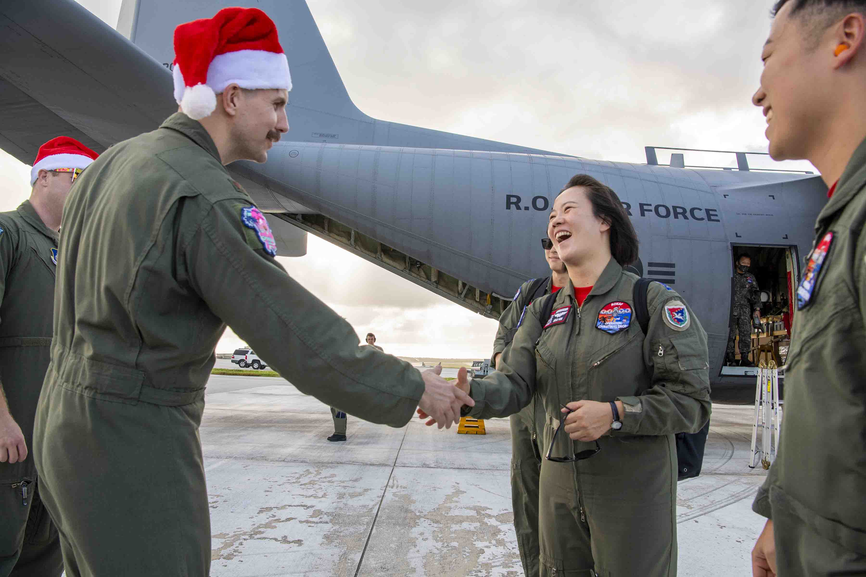 An airman in Santa hat shakes hands with a foreign service member in front of a plane parked on a flight line.