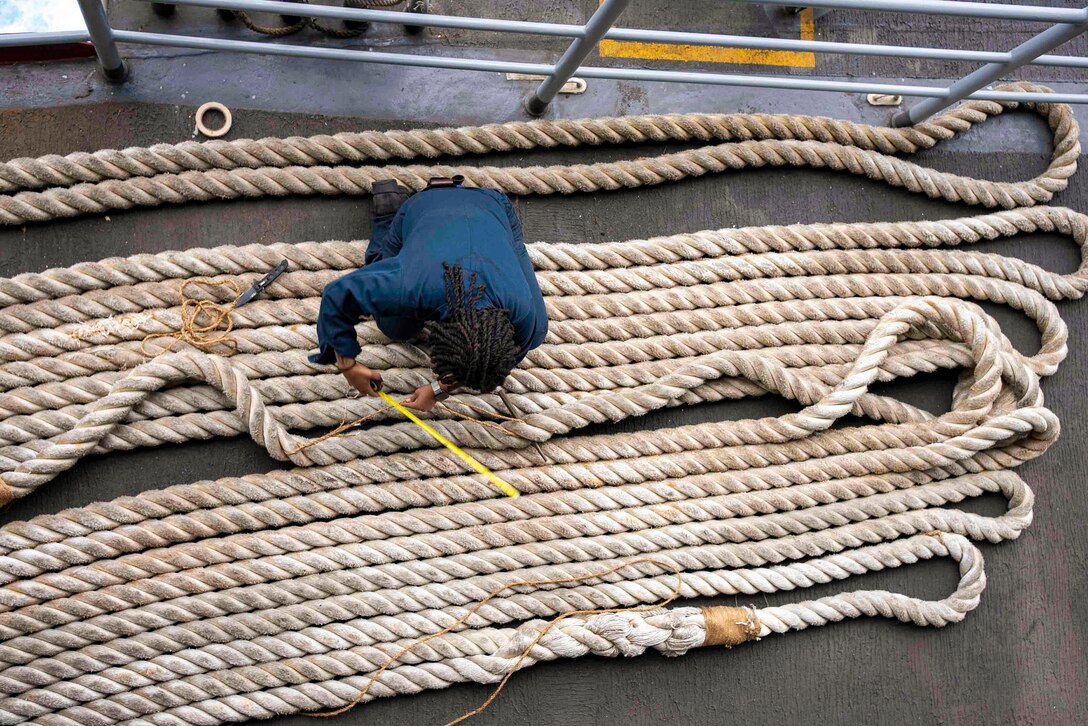 A sailor kneels on rows of rope aboard a ship while measuring string attached to the rope as seen from above.