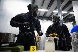 Two people in blue HAZMAT suits assess a jar full of a simulated suspicious liquid.