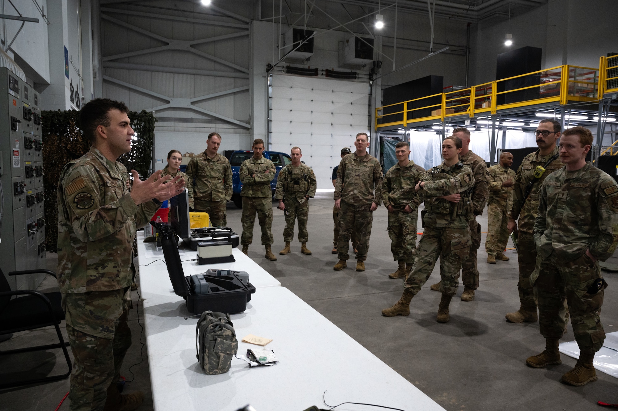 A uniformed military member, left, briefs a larger group of military personnel in a warehouse.