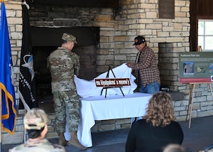 Colonel Mark Shoemaker stands on the left and Levi Zevalla stands on the right while they both unveil a street sign on top a table.