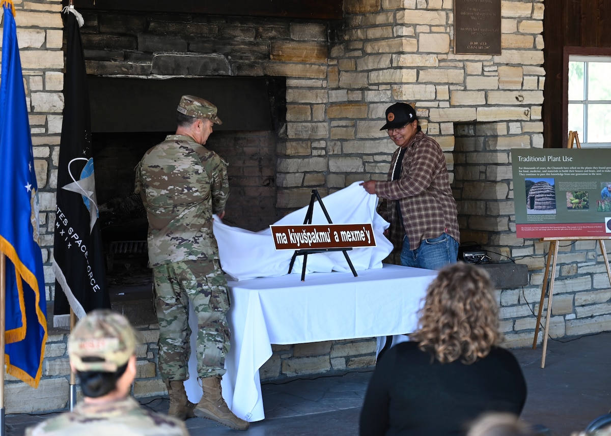 Colonel Mark Shoemaker stands on the left and Levi Zevalla stands on the right while they both unveil a street sign on top a table.