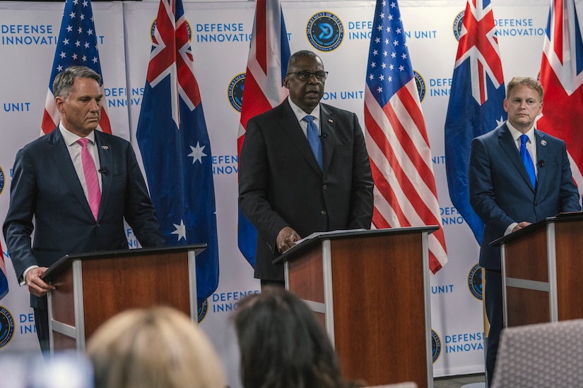 Three people stand behind podiums with flags behind them.