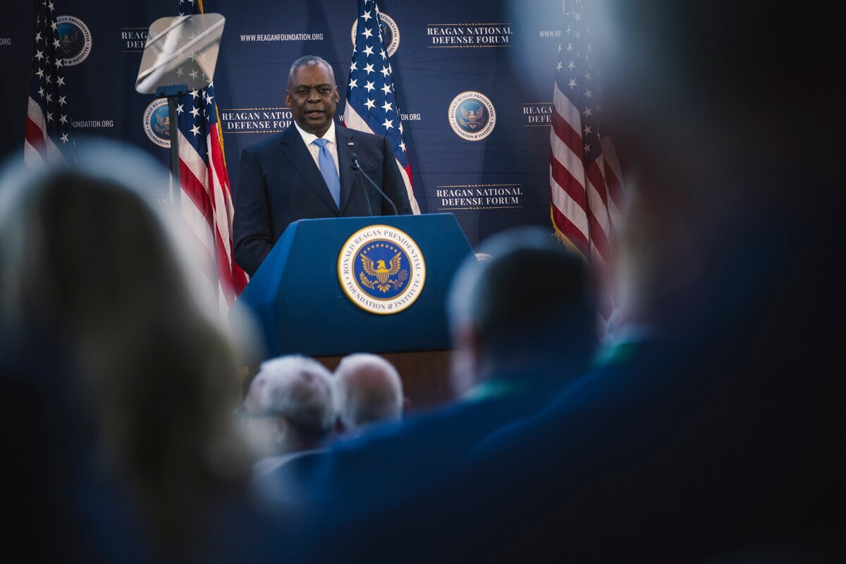 A group of people listen to a man speaking from a podium.