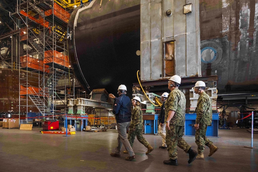 Service members in uniforms and wearing hard hats walk through an industrial facility.