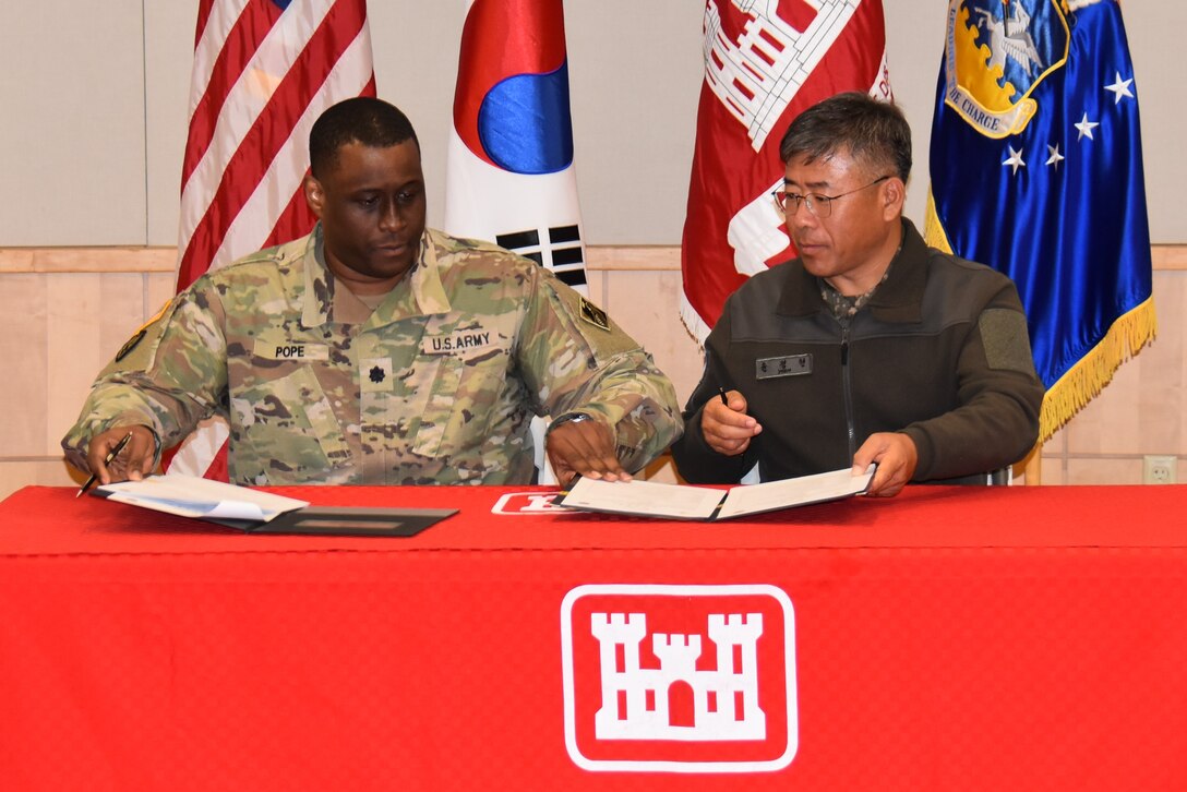 Lt. Col. Michael Pope, US Design and Construction Agent (DCA), and Lt. Col. Yun, Jeong Hyun, RoK DCA, sign transfer documentation in front of flags representing both countries.