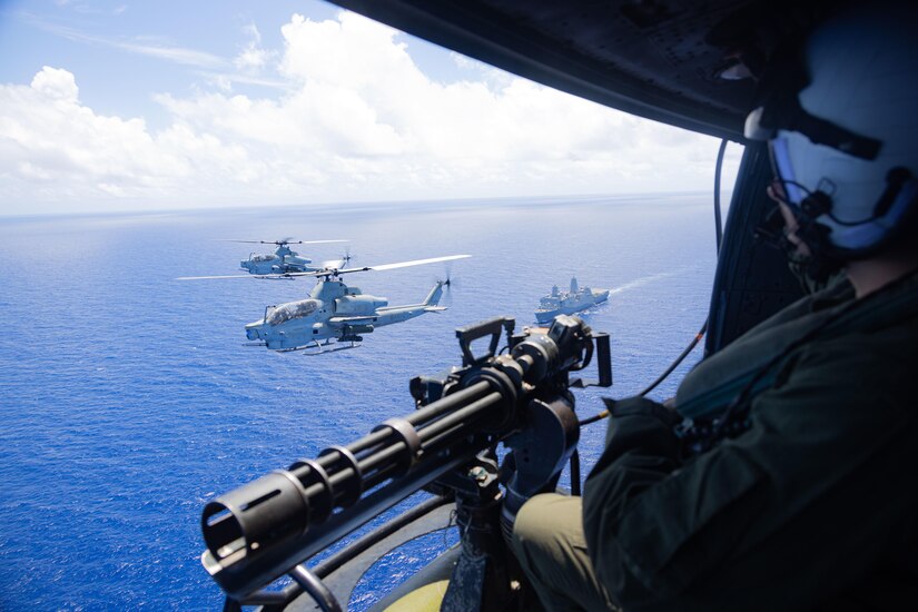 A service member operates a machine gun from a military aircraft flying over water as helicopters fly nearby and a ship sails below them.