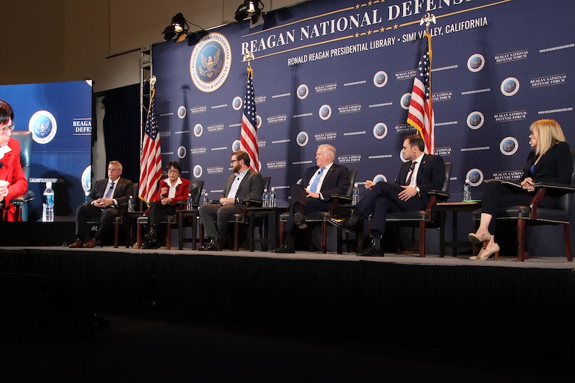 People sit on a stage during a panel discussion with American flags behind them.