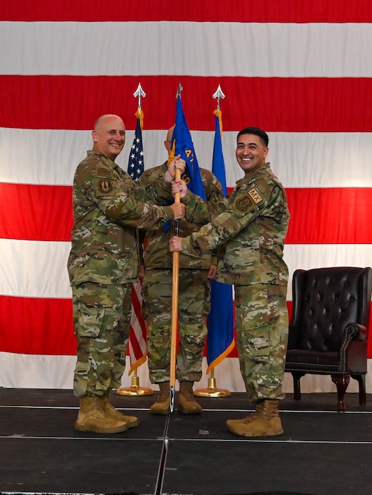 Two men in camouflage uniforms hold a blue flag between them in front of a very large American flag.