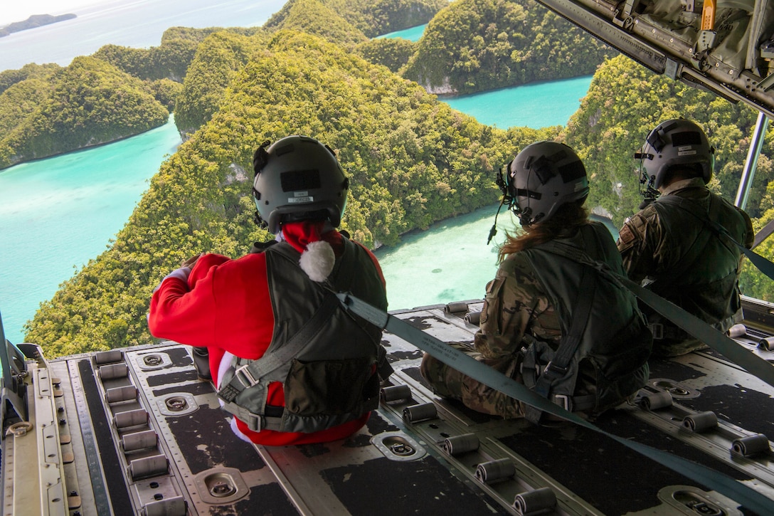 Three airmen, including one dressed as Santa, sit on the open ramp of an aircraft flying over islands.