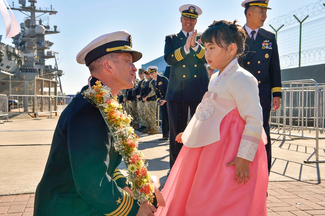 A sailor kneels while wearing lei and speaking to a child dressed in South Korean garb as fellow service members watch from the background.