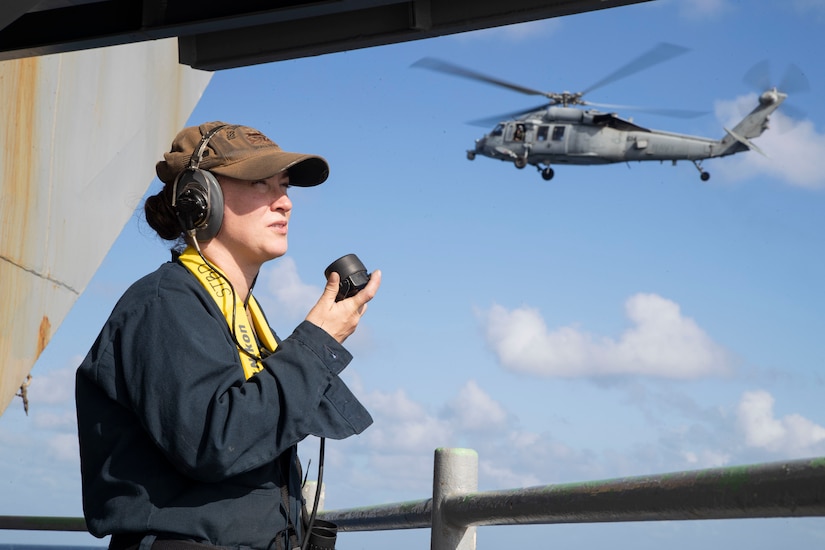 A sailor stands watch on a ship with a helicopter flying in the distance.