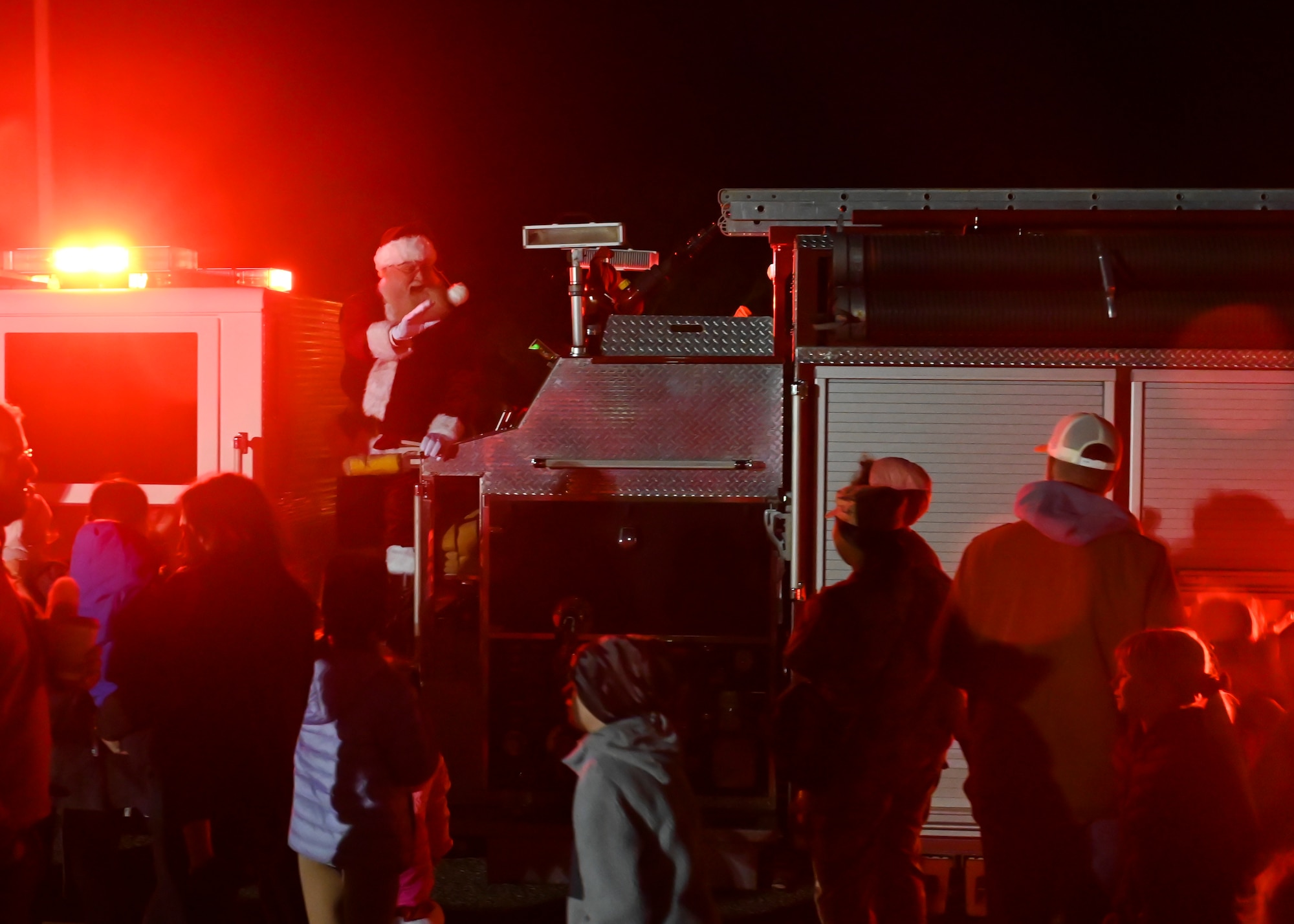 Santa Claus rides a fire truck next to a crowd of families.
