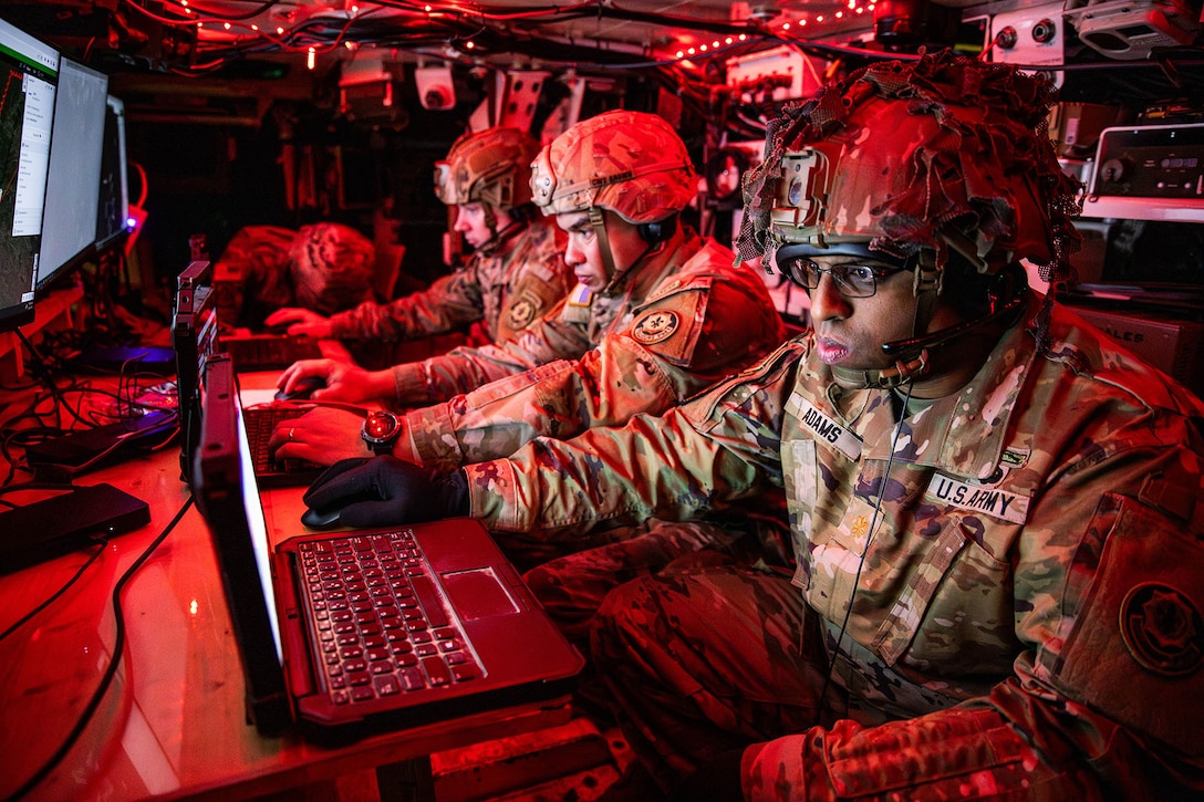 Three soldiers sit side by side while communicating in a Stryker armored vehicle.
