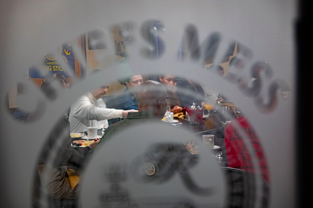 A group of service members are seen eating around a table through a glass door .