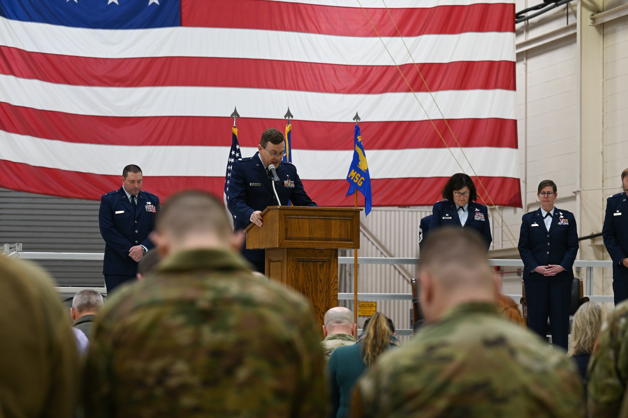 Lt. Col. Alex Jack, 434th Air Refueling Wing chaplain, leads the invocation during a change of command ceremony at Grissom Air Reserve Base. Jack is delivering the remarks at a podium on a stage with a large American flag in the background and several uniformed service members on stage behind him.
