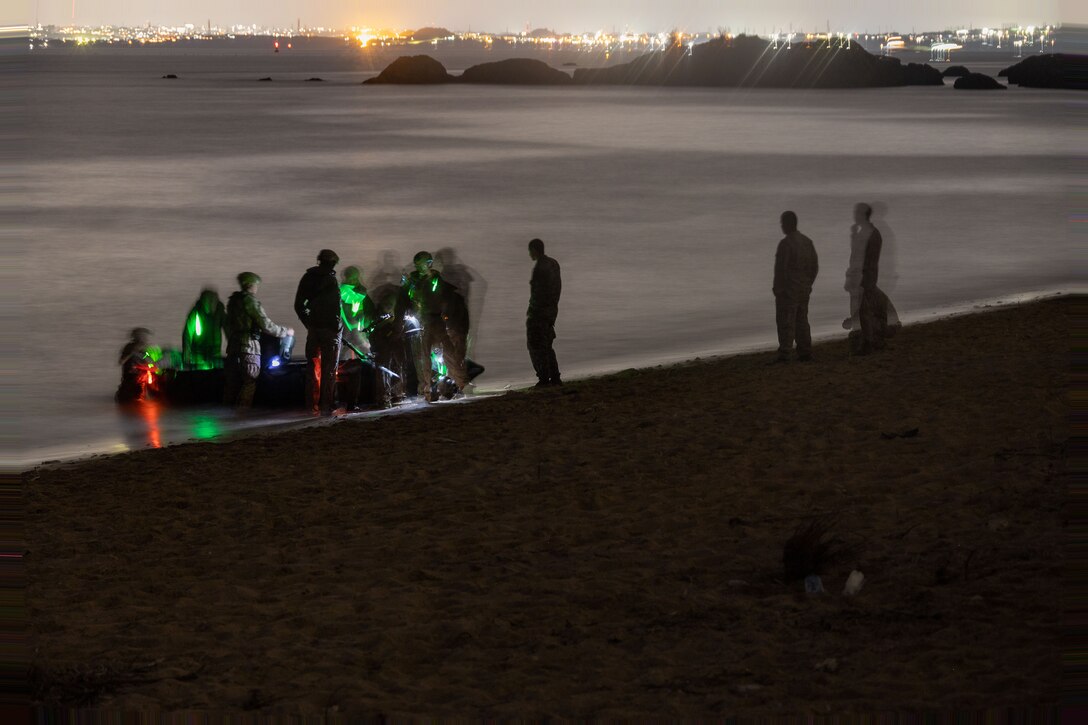 A group of Marines train on a rubber raiding craft on the shoreline as others watch at night. Lighting on the image is slightly blurred and city lights can be seen in the background.