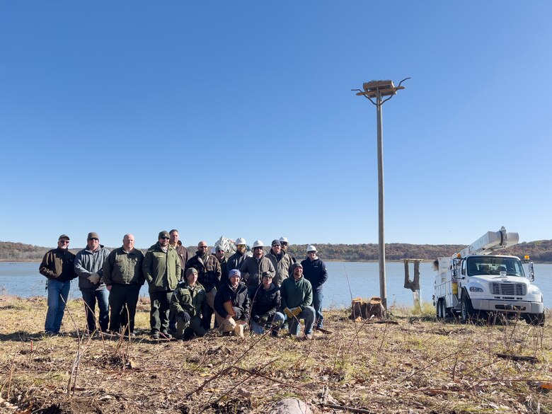 Several people stand and pose for a group photo to the left with a 40-foot tall pole and nest structure and a utility truck to the right.