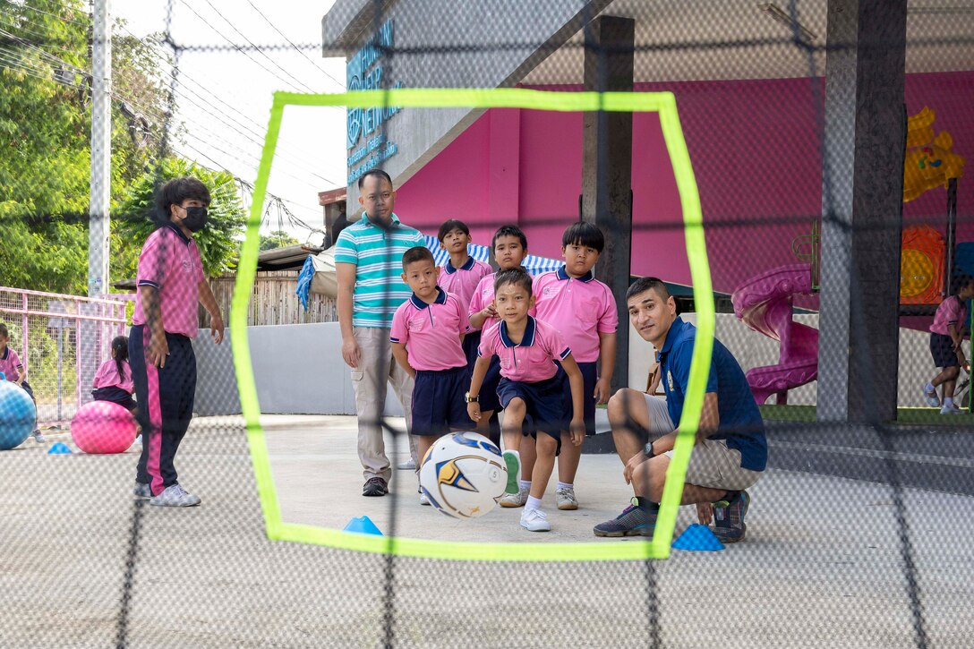 A sailor kneels next to a child as they kick a ball into a goal as seen through a hexagon cutout in the net while fellow children watch.